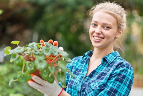 A young woman with blonde hair tied back, smiling and wearing a blue plaid shirt and gardening gloves, is holding a small potted plant with green leaves and reddish-brown stems. She is standing outdoors in a garden setting, with a background of lush green foliage and trees, suggesting a well-maintained backyard or front garden in Stoke Newington. The scene is bright, indicating a sunny day, and the woman appears to be engaged in gardening activities such as planting or tending to flower beds, which aligns with services offered by Landscapers Stoke Newington in the local area. The focus is on her cheerful expression and the healthy plant she holds, emphasizing the healthy growth of garden flora and the importance of attentive outdoor maintenance and landscaping care. This image demonstrates the type of gardening service that enhances outdoor spaces in residential environments within the local postcode region.