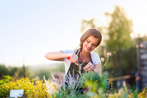 A woman with light brown hair, smiling while tending to a flower bed in an outdoor garden setting. She is wearing a light blue top and is using a small gardening tool to work among a variety of plants, including flowering plants and green foliage. The garden features a mix of blooming flowers and lush green shrubs with a neatly maintained lawn in the foreground. In the background, there are trees and a clear sky, suggesting a sunny day with natural daylight illuminating the scene. The setting appears to be a well-kept backyard or front garden designed for leisure and gardening, with a focus on floral and plant maintenance, reflecting typical features found in residential gardens in Stoke Newington. This image emphasizes outdoor care and gardening activities suitable for professional landscaping services like those offered by Landscapers Stoke Newington.