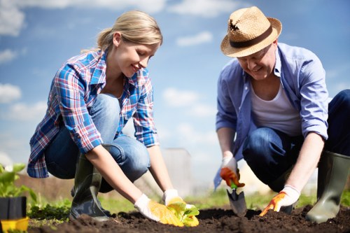 The image depicts two individuals working together in a garden, with a young woman and an older woman planting or tending to soil in a landscaped outdoor space. The young woman, dressed in a checked shirt and jeans, is kneeling on a patch of soil, carefully handling a small plant or seedling. The older woman, wearing a wide-brimmed hat, a blue shirt, and jeans, is also crouched down, using a trowel or gardening fork to work the soil. The garden features a well-maintained lawn area with lush, dense grass surrounding the planting beds, which are bordered by soil that appears freshly tilled. In the background, there is a clear sky with some clouds, suggesting pleasant weather. The scene conveys active gardening, focusing on planting and soil preparation in a tidy, thoughtfully landscaped outdoor area within Stoke Newington, reflecting professional gardening and landscaping services by Landscapers Stoke Newington.