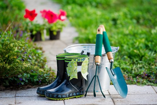 A pair of dark blue rubber gardening boots with a camouflage pattern, placed on a paved surface in a garden setting, with a garden trowel and hand fork leaning against a metal bucket containing gardening tools, surrounded by green plants and flowering potted plants in the background. The scene suggests outdoor gardening activities typical of a well-maintained Stoke Newington garden, with lush greenery, flowering plants, and a neat arrangement of gardening equipment, indicating ongoing gardening or landscaping work funded by local services. The natural outdoor environment features a mix of grass and plants, with sunlight providing bright, natural lighting, highlighting the vibrant colours of the flowers and the condition of the garden tools, aligning with gardening and lawn care themes relevant to the Stoke Newington area.