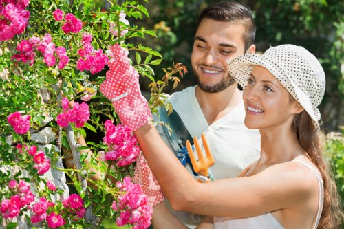 A man and a woman are engaged in gardening activities in a well-maintained outdoor garden space. The woman is wearing a wide-brimmed hat and pink gardening gloves, actively trimming a flowering shrub with vibrant pink blossoms. The man, standing close beside her, is smiling and holding a small garden fork, observing her work. The garden features a lush green lawn in the foreground, bordered by neatly maintained flower beds and a hedge line in the background. Tall trees and dense foliage provide a natural backdrop, suggesting a peaceful, mature outdoor area typical of residential gardens in the Stoke Newington area. The scene is illuminated by natural daylight, with soft, diffuse sunlight filtering through the tree canopy, highlighting the vivid colors of the flowers and greenery. This image reflects professional gardening and landscaping efforts focused on cultivating a healthy, visually appealing garden environment, supported by local gardening services such as those provided by Landscapers Stoke Newington.