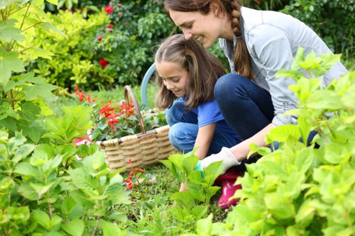 In a lush, well-maintained garden, a woman and a young girl are engaged in outdoor gardening activities. They are surrounded by a variety of green plants, shrubs, and flowering bushes, with some vibrant red flowers visible in the background. The girl is crouched down, holding a small gardening basket and appears to be planting or tending to the soil, while the woman is smiling and leaning over, guiding or assisting her. The garden features a mix of grass, soil beds, and dense foliage, with a natural, slightly wild appearance that suggests ongoing gardening work. The lighting indicates a bright, clear day, with natural sunlight illuminating the scene. This setting is typical of a suburban front or back garden in Stoke Newington, reflecting a focus on gardening and outdoor maintenance which local landscaping services might support, emphasizing natural plant growth and garden nurturing.