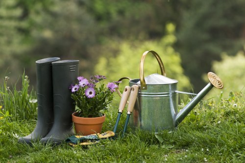 The image depicts a neatly maintained garden with a lush, green lawn in the foreground. On the grass, there are a pair of black rubber gardening boots standing upright, next to a small flowerpot filled with vibrant purple and white flowers. Nearby, several gardening hand tools such as a trowel and pruning shears are arranged on the ground. A metallic watering can with a wooden handle and a long spout is prominently placed beside the tools, ready for watering plants. The background features blurred greenery, including trees and shrubs, characteristic of a well-kept outdoor space in Stoke Newington, suggesting a calm and inviting garden setting suitable for planting and outdoor maintenance activities. This scene reflects typical gardening elements and tools used for planting services offered by Landscapers Stoke Newington, showcasing a prepared and lush environment ideal for garden care in the local area, with natural lighting indicating a bright, slightly overcast day.