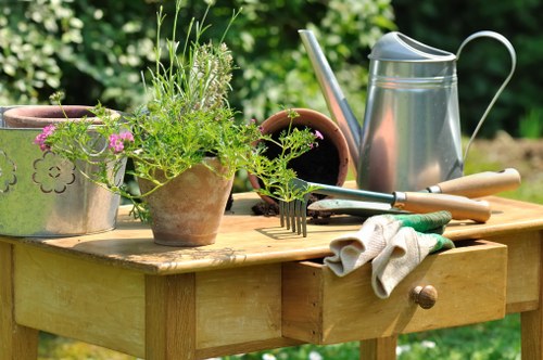 A wooden garden table in an outdoor setting with various gardening tools and containers arranged on its surface. The table holds a terracotta pot filled with a mix of green plants and herbs, some with purple flowers, alongside a metal watering can with a curved handle and spout. There is a small open terracotta pot, a pair of gardening gloves resting on the edge of the table, and a hand rake with green handle. In the background, blurred lush green foliage and trees suggest a thriving garden environment, providing a natural and inviting atmosphere typical of outdoor spaces in Stoke Newington. The scene appears to be set on a bright, clear day, suitable for gardening activities supported by local garden maintenance services.