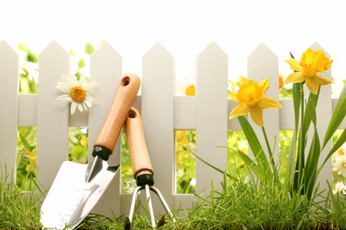 A close-up view of a domestic garden featuring a white picket fence in the background, with yellow daffodils and white daisies growing alongside it. In the foreground, there is a patch of lush green grass with a set of gardening tools, including a trowel and a hand rake with wooden handles, leaning against the fence. Behind the tools, soil beds are visible, suggesting ongoing planting or garden maintenance. The garden appears well-maintained, with natural light illuminating the scene, indicating a clear day. This outdoor space is part of a residential garden, showcasing typical elements of spring planting and lawn care, suitable for gardening services offered by companies like Landscapers Stoke Newington, serving the N16 postcode area in London.