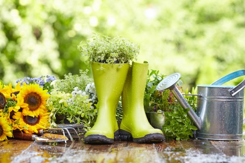 A garden scene featuring a weathered wooden table with a variety of gardening tools and decorative elements. In the center, a pair of bright green rubber gardening boots are repurposed as plant holders, filled with small white flowering plants. To the left, a small bouquet of vibrant yellow sunflowers and other mixed flowers rests on the table, adding natural colour contrast. On the right side, a silver metal watering can with a blue handle is positioned beside additional potted greenery, including leafy plants in small containers. The background displays a lush, softly blurred garden with green foliage, suggesting an outdoor space in spring or summer. The scene is well lit with natural sunlight, highlighting the textures of the wooden surface, plant leaves, and the reflective metal of the watering can. This arrangement emphasizes outdoor gardening activities suitable for a garden or backyard in Stoke Newington, with a focus on plant care and garden decoration reflective of professional gardening services by Landscapers Stoke Newington.