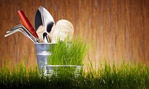 The image shows a close-up view of gardening tools and supplies placed in a small metal bucket, situated outdoors on a lush, green grass lawn. The tools include a red-handled rake, a metal trowel, and a hand rake, with a ball of natural twine positioned among them. The background features a wooden surface, possibly part of a garden shed or fencing, suggesting a garden setting in Stoke Newington. The natural lighting highlights the vibrant green grass and the textures of the metal tools, conveying an active outdoor gardening environment. The scene reflects essential gardening activities, emphasizing outdoor maintenance and planting projects that landscapers like Landscapers Stoke Newington might undertake, aligning with their focus on sustainable and environmentally conscious garden care in the local area.