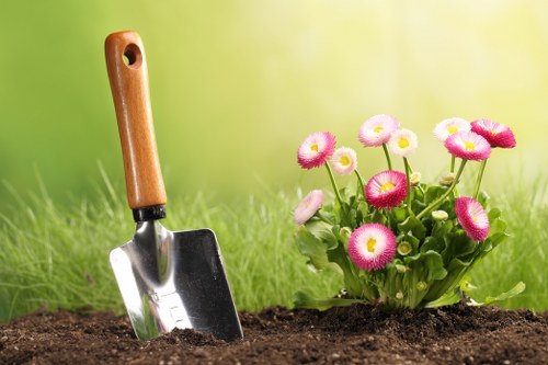 A close-up view of a garden bed with freshly turned dark brown soil, featuring a small hand trowel with a wooden handle partially inserted into the ground, positioned next to a cluster of vibrant pink and white daisy-like flowers with green foliage. The background consists of a softly blurred green lawn and trees, suggesting a well-maintained outdoor space typical of residential gardens in Stoke Newington. The scene is lit by natural daylight, indicating mild weather conditions, and illustrates a setting suitable for gardening and outdoor maintenance services provided by Landscapers Stoke Newington, emphasizing attention to flower bed preparation and plant care.