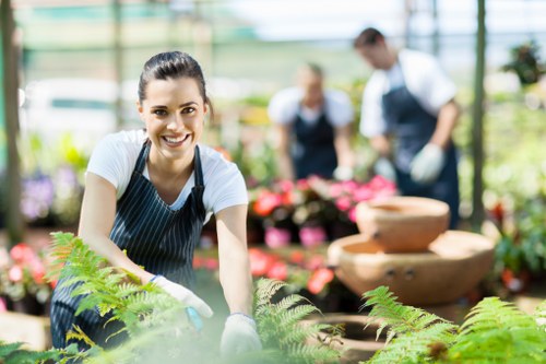 A smiling woman wearing a striped apron and gardening gloves is kneeling in a greenhouse with a lush green garden foreground, showcasing vibrant ferns and leafy plants. Behind her, two other gardeners in aprons are tending to a variety of potted flowers and plants, with pink blossoms visible. The greenhouse structure features large glass panels allowing natural light to illuminate the scene. The garden area includes soil beds, paved pathways, and decorative stone water features in the background, all within a well-maintained outdoor space typical of professional gardening services in Stoke Newington. The environment appears healthy and vibrant, with an emphasis on thriving greenery and organized planting arrangements, reflecting the high standards of landscaping and garden care. The weather is bright and clear, supporting effective plant growth, while the scene captures a professional, collaborative effort in outdoor garden maintenance, highlighting the expertise of Landscapers Stoke Newington in creating beautiful, sustainable landscaped environments within London.