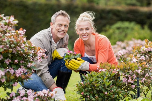 A smiling middle-aged man and woman are working together in a well-maintained garden in Stoke Newington, surrounded by lush green grass, flowering bushes with pink and white blossoms, and neatly arranged plants. The man is dressed in a light grey shirt and jeans, while the woman wears an orange cardigan and pink gardening gloves. They are kneeling on the lawn, holding a small plant between them, with gardening tools nearby. The background features mature trees and a softly blurred garden landscape, indicating a sunny day with natural light illuminating the scene. The garden appears to be carefully landscaped, with a mix of lawn, flowering shrubs, and mulched flower beds, reflecting professional outdoor maintenance and gardening expertise common in the area.